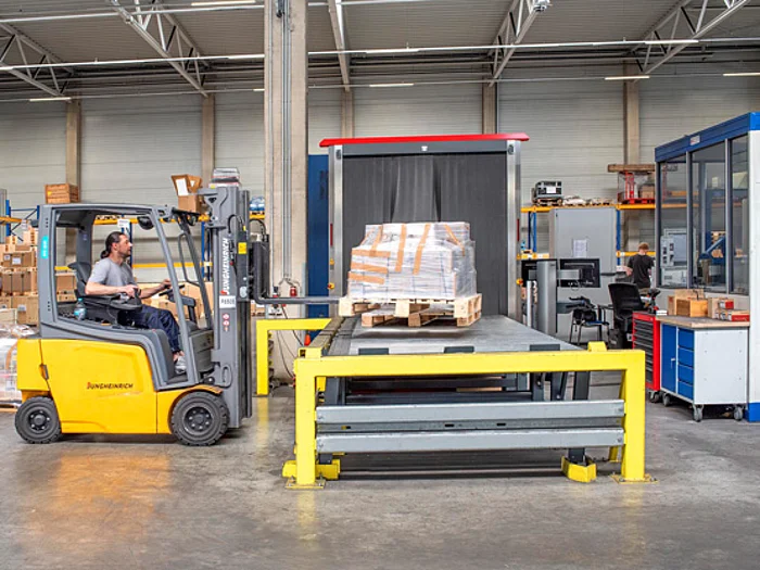 In einer Industriehalle transportiert ein Gabelstapler eine Palette mit Kartons zu einer Maschine. Die Palette steht auf einer gelben Plattform vor einer Frachtröntgenanlage mit transparentem Vorhang. Weitere Maschinen und Regale befinden sich im Hintergrund.