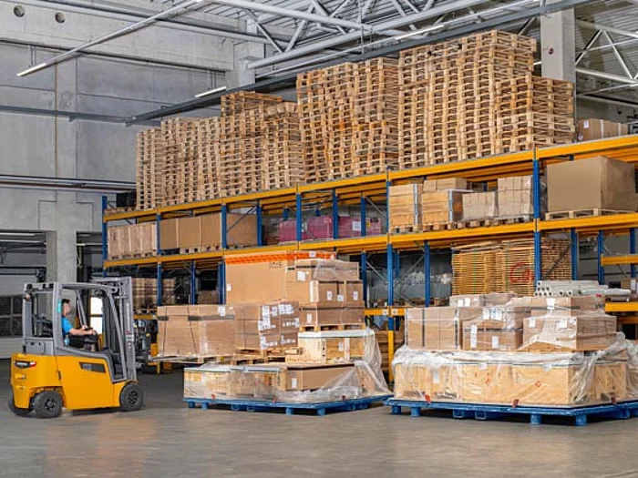 In einer Lagerhalle stehen mehrere Paletten mit verpackten Kartons auf dem Boden. Ein Gabelstapler fährt vor hohen Regalen mit gestapelten Holzpaletten und Kartons entlang. Die Regale sind mehrstöckig und aus Metall.