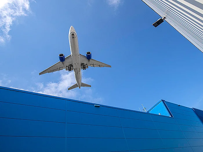 Ein Flugzeug ist von unten vor blauem Himmel zu sehen. Im Vordergrund befindet sich die Fassade eines blauen Industriegebäudes. Das Flugzeug fliegt über das Gebäude hinweg.